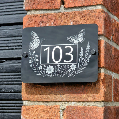 Decorative house number sign with butterflies and flowers on a brick wall.
