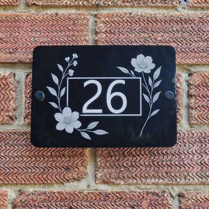 House number sign with floral side blooms design on a black acrylic against a brick wall.